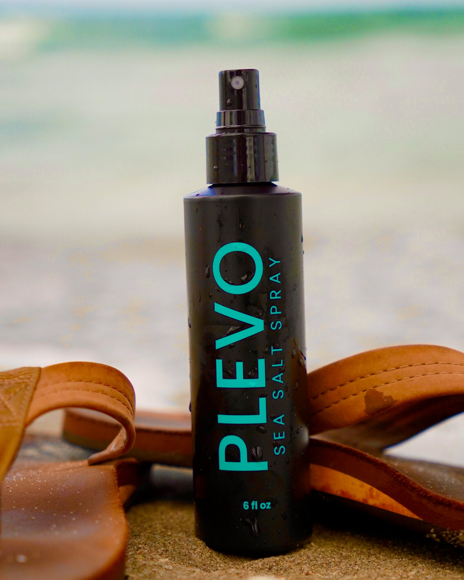 The PLEVO sea salt spray bottle standing in wet sand beside a pair of leather sandals, ocean behind